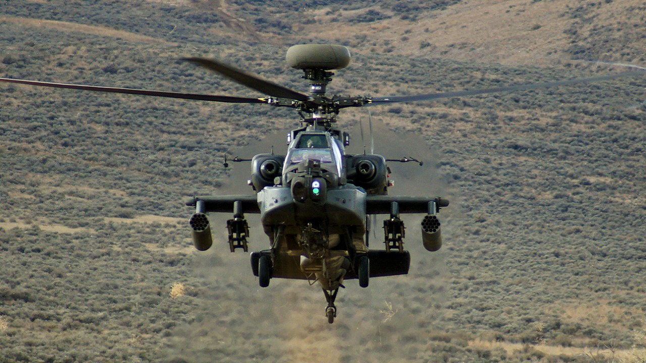 An AH-64E Apache helicopter flying toward the camera over a hillside