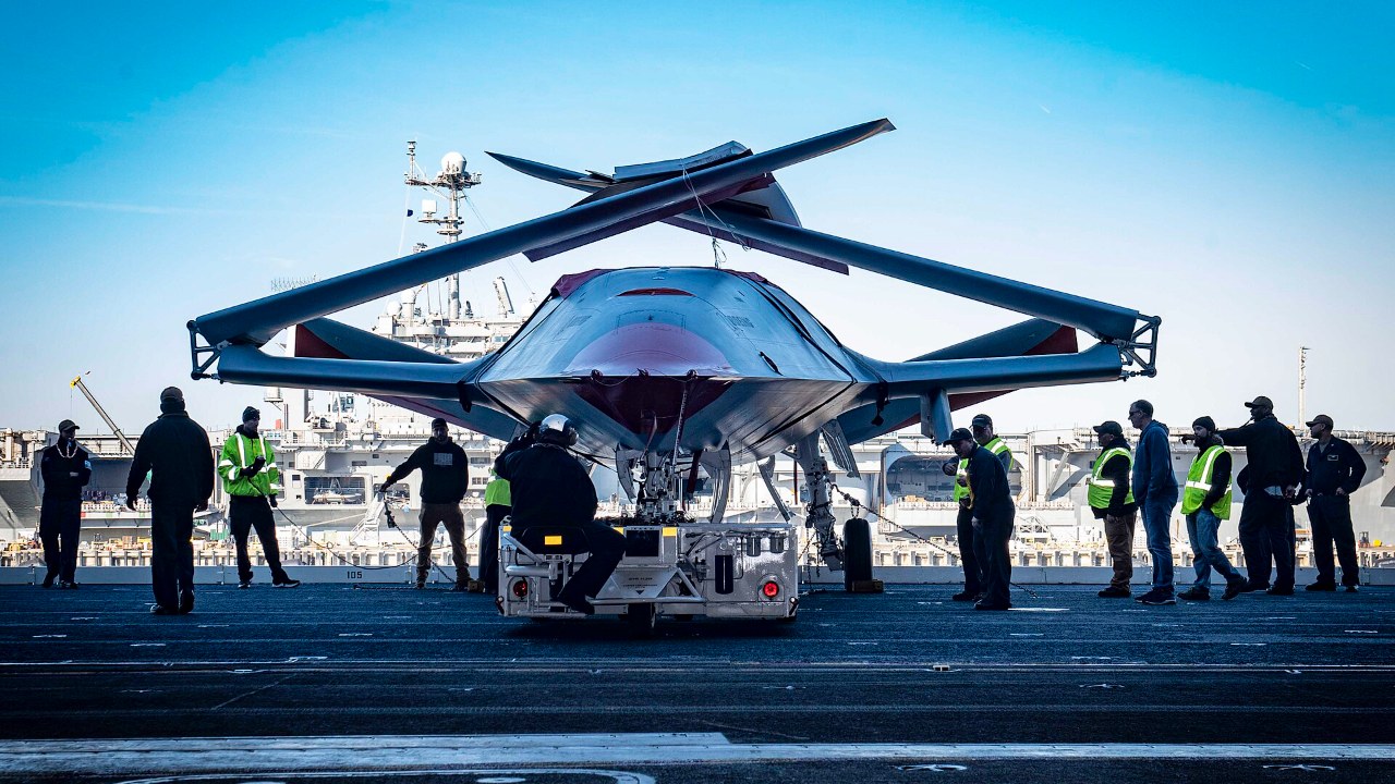 The MQ-25 unmanned aircraft with folded wings on a carrier deck surrounded by sailors
